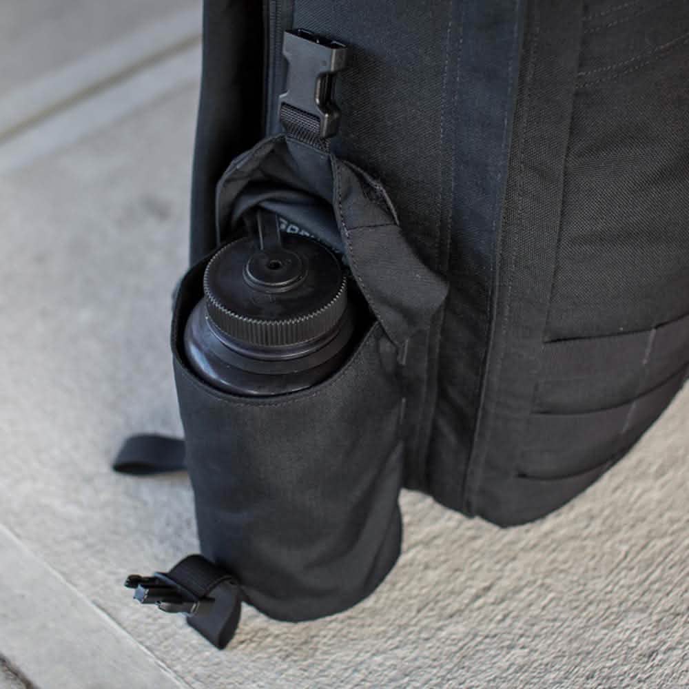 A close-up of a black backpack by GORUCK features the Full Panel Water Bottle Pocket, securely holding a Nalgene-style water bottle in its side pocket. The backpack sits on a concrete surface, highlighting its practicality with the water bottle's screw-top lid for easy and accessible hydration.