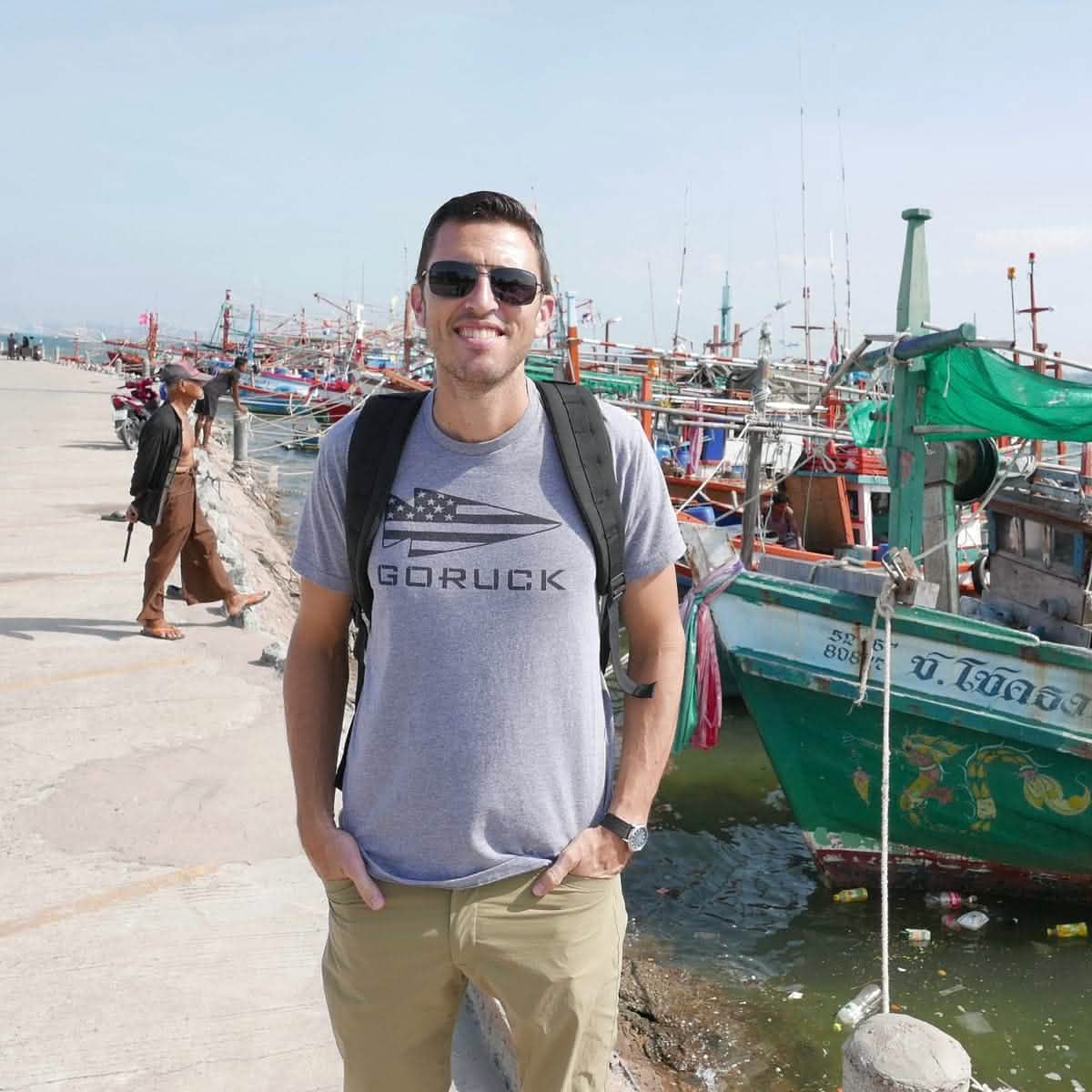 A man stands smiling on a dock, wearing the GORUCK USA Tee - Tri-blend by vendor-unknown, with colorful fishing boats in the background.