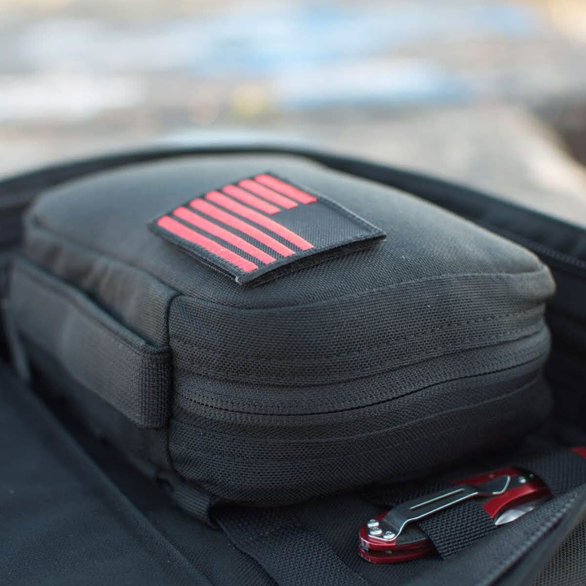A close-up of the GR1 Field Pocket by GORUCK, a black electronics organizer with a red-striped patch, placed on top of a black bag. A red and silver tool with a clip is visible beside the pouch in an outdoor setting.