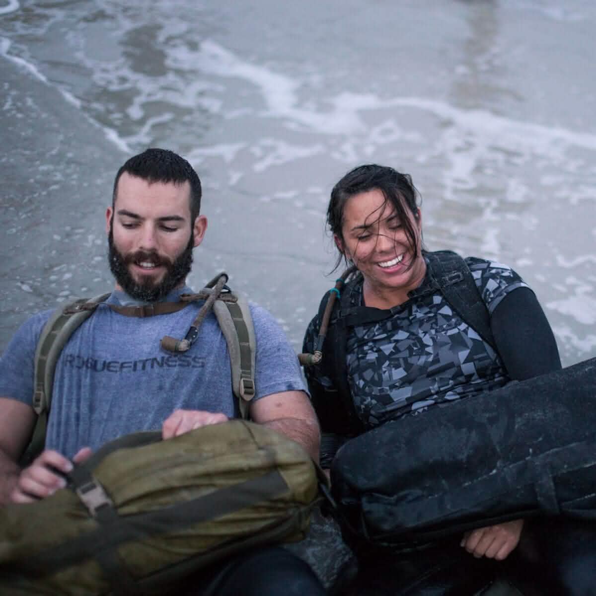A man and a woman in wet clothing sit on a sandy beach, smiling. They each hold backpacks equipped with a GORUCK Hydration Valve Mouthpiece and wear military gear. The ocean waves gently touch them in the background, adding to their adventurous spirit.