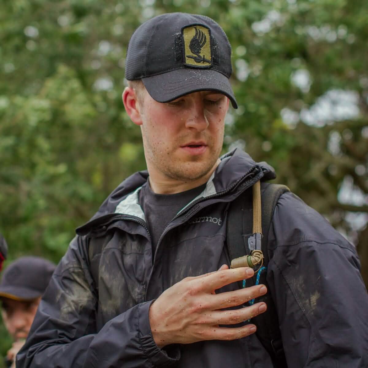 A man wearing a black cap and jacket examines his mud-splattered hand while standing outdoors with greenery behind him. He is holding a GORUCK Hydration Valve Mouthpiece attached to his backpack strap. Another person is partially visible in the background.