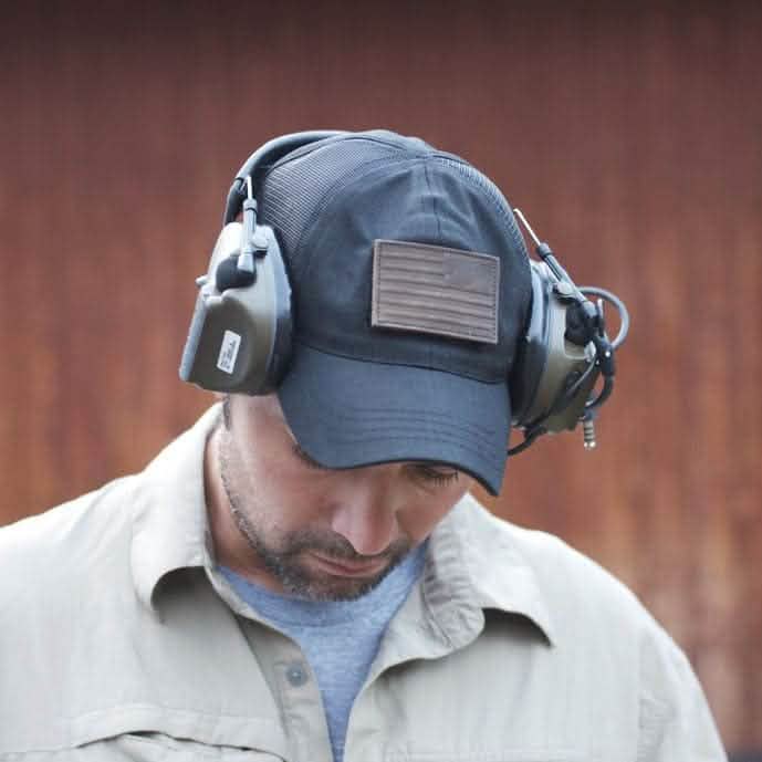A person wearing a black cap featuring a leather reverse flag patch from vendor-unknown and large noise-canceling headphones attached using a VELCRO® backing. The person is looking down, clad in a beige jacket over a gray shirt, with the background appearing blurred and reddish-brown.