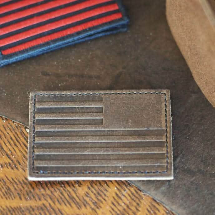 The Patch - Leather Reverse Flag, crafted by vendor-unknown from full grain cowhide leather, features a flag design with horizontal stripes and a blank square in the top left corner. It is placed on a wooden surface next to another fabric patch that shares a similar red and black pattern.