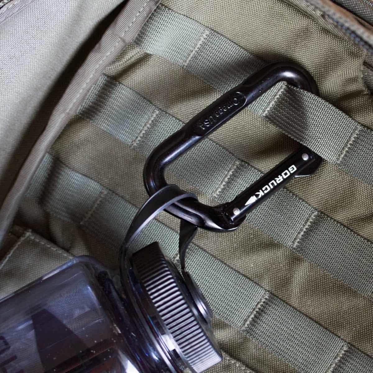 Black GORUCK branded carabiner attached to olive green ruck sack webbing with a black water bottle nearby