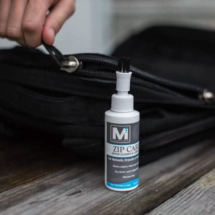 Using a bottle equipped with an attached brush, someone is applying Gear Aid's Zip Care - Zipper Cleaner & Lubricant to the zipper of a black bag. This product ensures smooth operation and rests on a wooden surface, making it an ideal preparation for GORUCK Events.