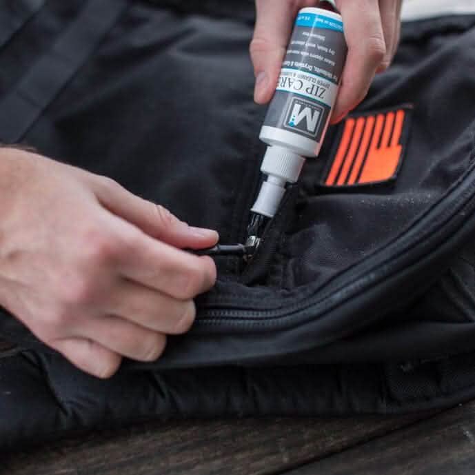 Close-up of a person carefully applying Gear Aid's Zip Care - Zipper Cleaner & Lubricant to a black zipper on a bag. The individual holds the zipper and tube gently over a wooden surface, preparing for GORUCK Events.