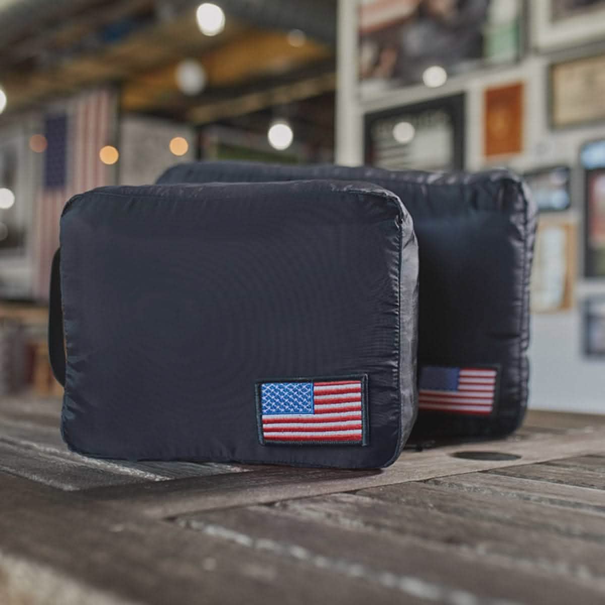 Two ultra-light nylon packing cubes, showcasing American flag patches, sit on a wooden table. In the softly blurred background of this indoor space are American flags and framed items—perfect travel companions from GORUCK for those seeking style and patriotism.