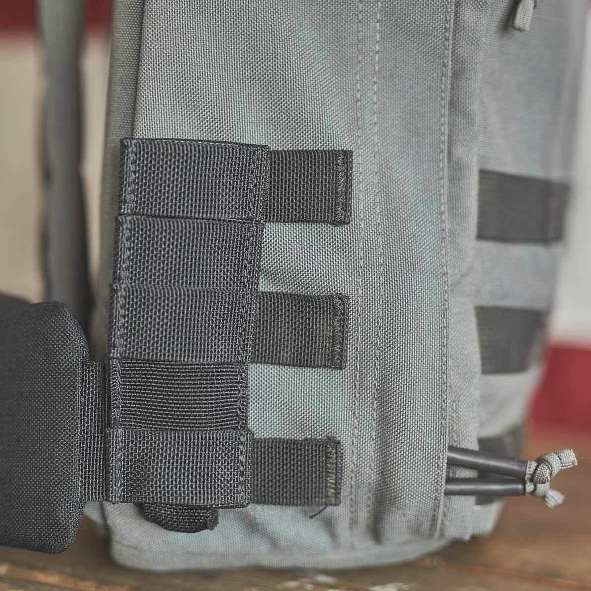 Close-up of the GORUCK backpack featuring a gray design with black straps, MOLLE webbing on the side, and its signature Padded Hip Belt for comfortable hip carry. Ideal for heavy loads, this sturdy backpack is displayed on a wooden surface with a blurred backdrop.