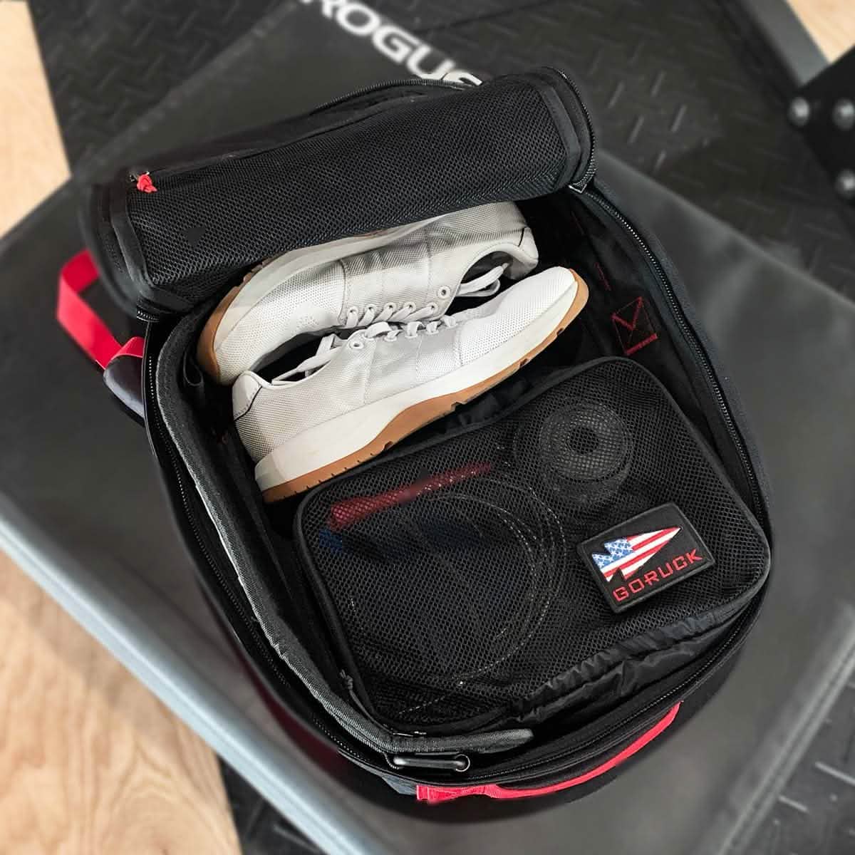 A Packing Cube from GORUCK, made of durable 200D nylon, holds white sneakers, a rolled-up towel, and a small pouch adorned with a GORUCK patch. Ideal for your travel ruck adventures, this cube keeps everything organized as it rests on the black mat.