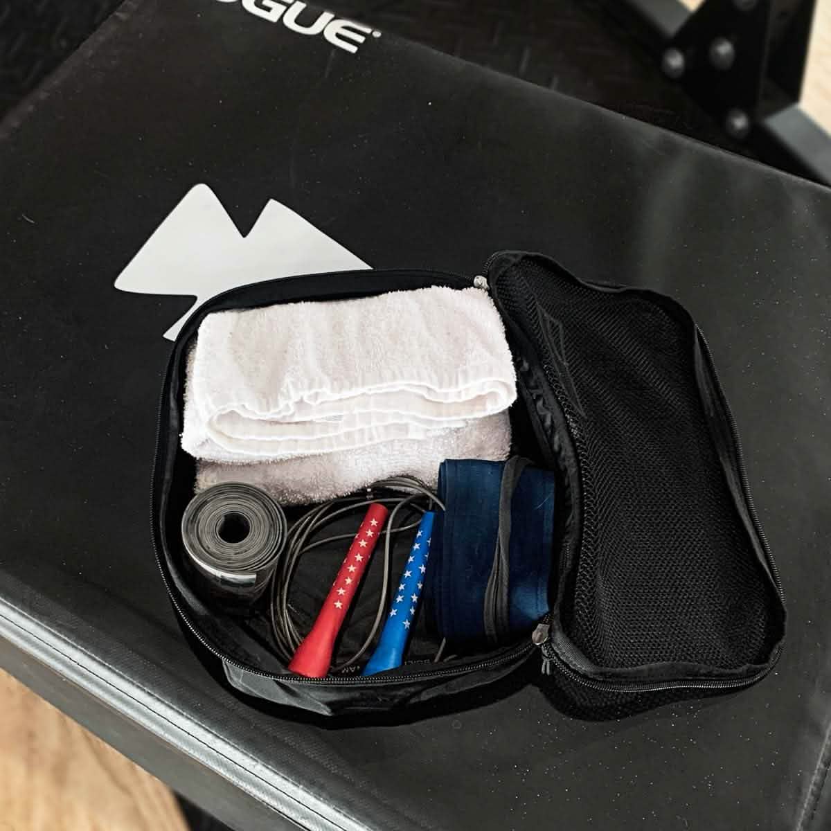 A black gym bag crafted from ultra-light nylon sits on a workout bench. Inside, you'll find a white towel, GORUCK Packing Cubes containing a roll of athletic tape, a jump rope with red and blue handles, and a resistance band—making it an ideal travel companion for maintaining your fitness routine on the go.