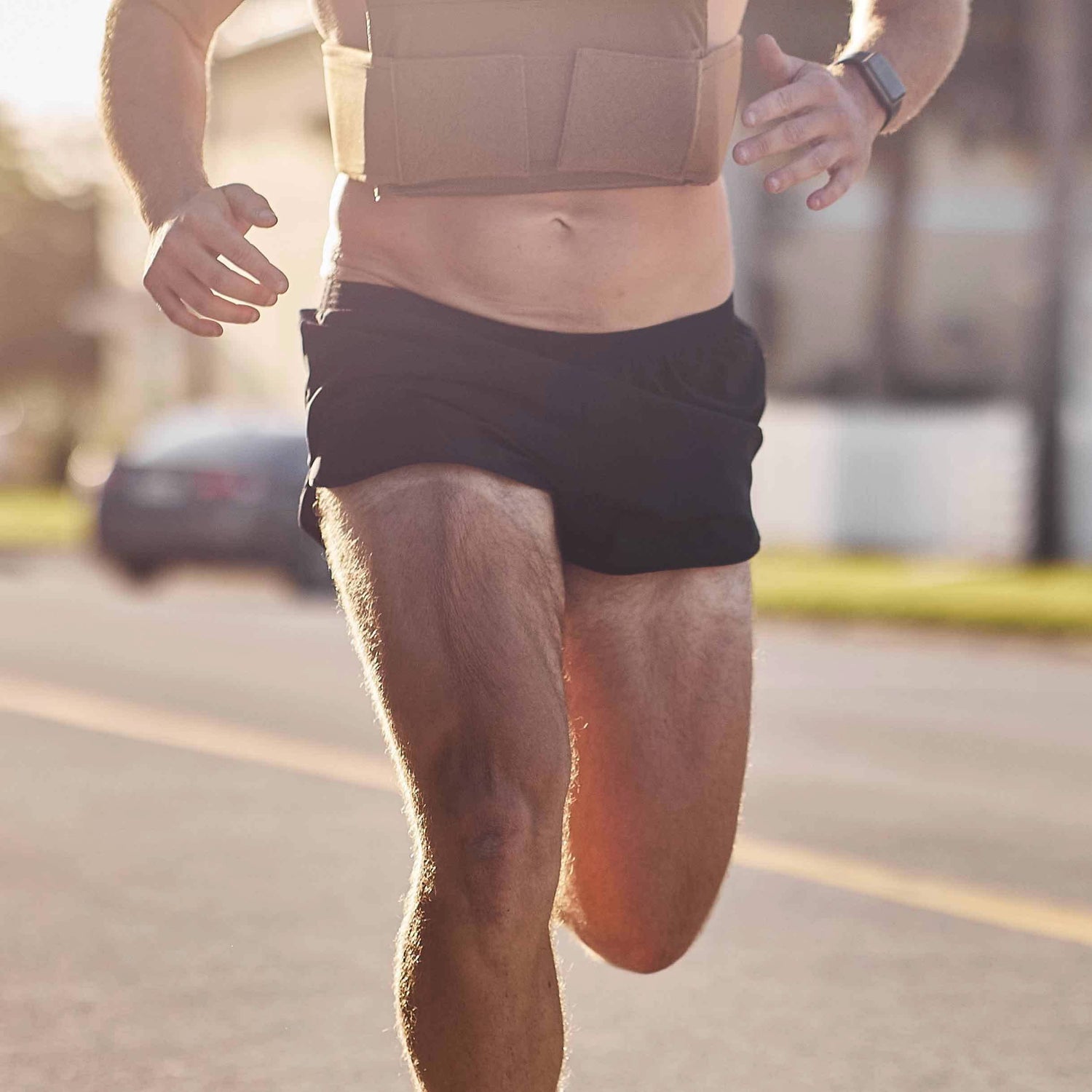 A person wearing Men’s Ranger Panties - ToughStretch by GORUCK and a vest is running on a sunlit road. They have a watch on their wrist, and the background is slightly blurred, showing a parked car and some trees. The shorts, made from ToughStretch fabric, provide ultimate comfort during their energetic stride.