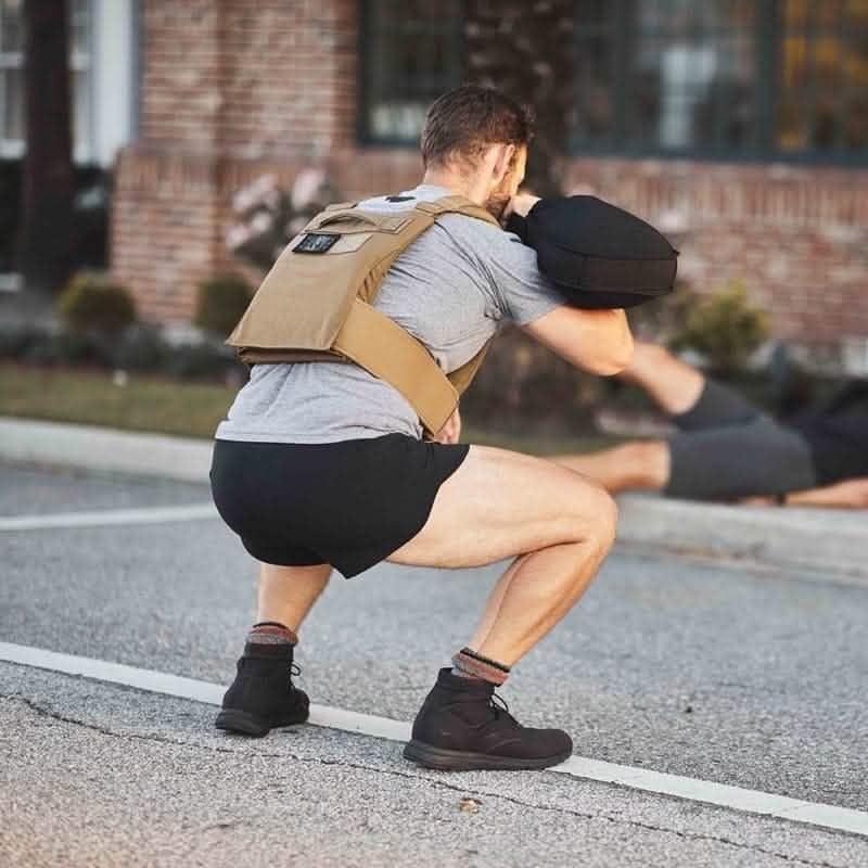 Man squatting outdoors wearing black shorts, gray shirt, tactical rucking weight vest, and black boots