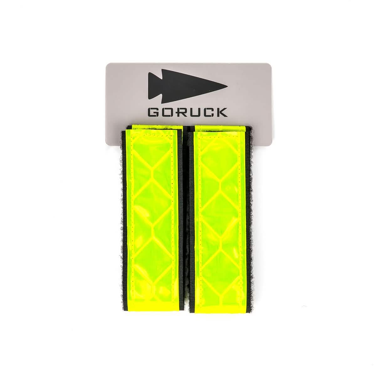 A pair of GORUCK Reflective Ruck Bands in bright yellow, mounted on a gray backing with an arrowhead symbol in black as part of the GORUCK logo. These straps are crafted to seamlessly integrate with MOLLE webbing and feature a diamond pattern to boost visibility and safety during nighttime use.