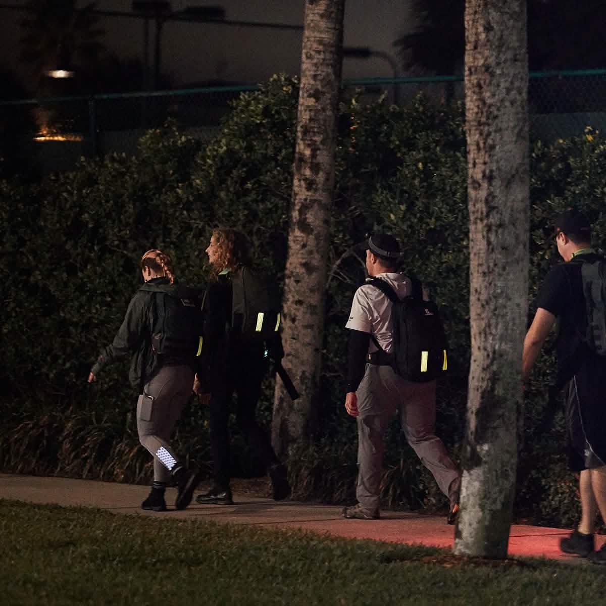 A group of four individuals strolls along a sidewalk at night, each with backpacks equipped with GORUCK Reflective Ruck Bands. The area is surrounded by trees and foliage, dimly illuminated by streetlights to ensure maximum visibility in the dark.