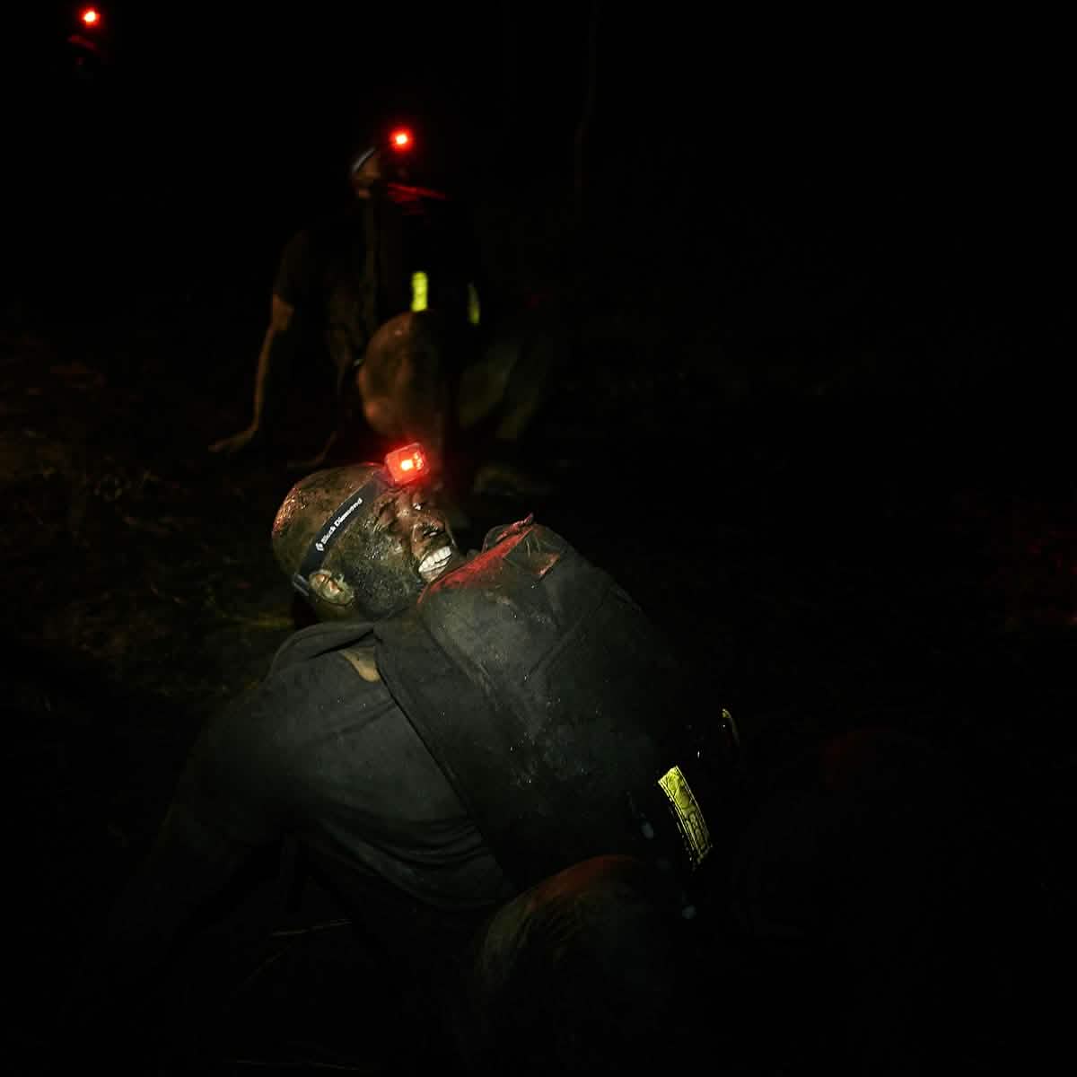 Three individuals are involved in a nighttime outdoor activity, each carrying backpacks equipped with GORUCK Reflective Ruck Bands and headlamps emitting red lights for improved visibility. The setting seems demanding and rugged, possibly suggesting an adventure race or survival training scenario.