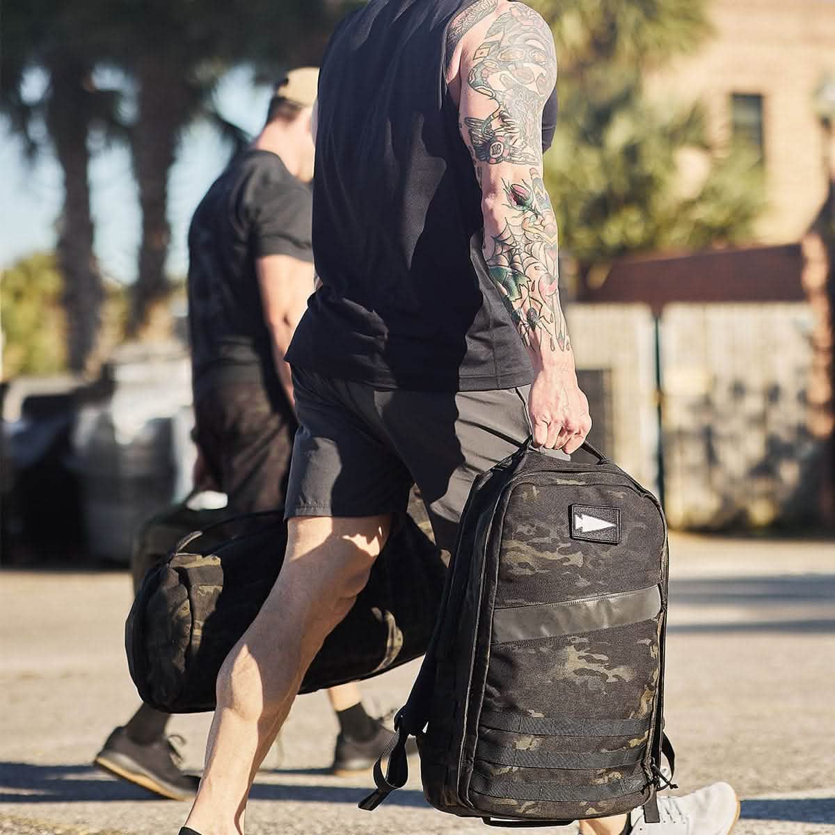 Two individuals are strolling outside, each carrying sizeable GORUCK backpacks adorned with embroidered Reflective Spearhead patches and dressed in casual athletic attire. One person sports full-arm tattoos. Behind them, palm trees mingle with a brick building against a backdrop of clear skies.
