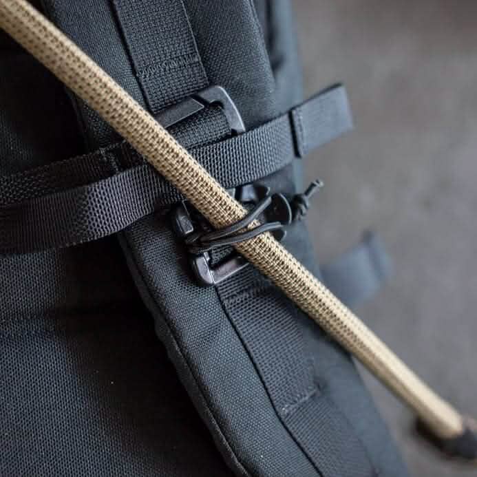 Close-up of a GORUCK black backpack featuring a tan walking stick secured to the side with an elastic shock cord and a Bladder Hose Retainer clip. The image highlights the durable fabric and fastening system, perfect for enthusiasts seeking reliability and style during GORUCK Events.