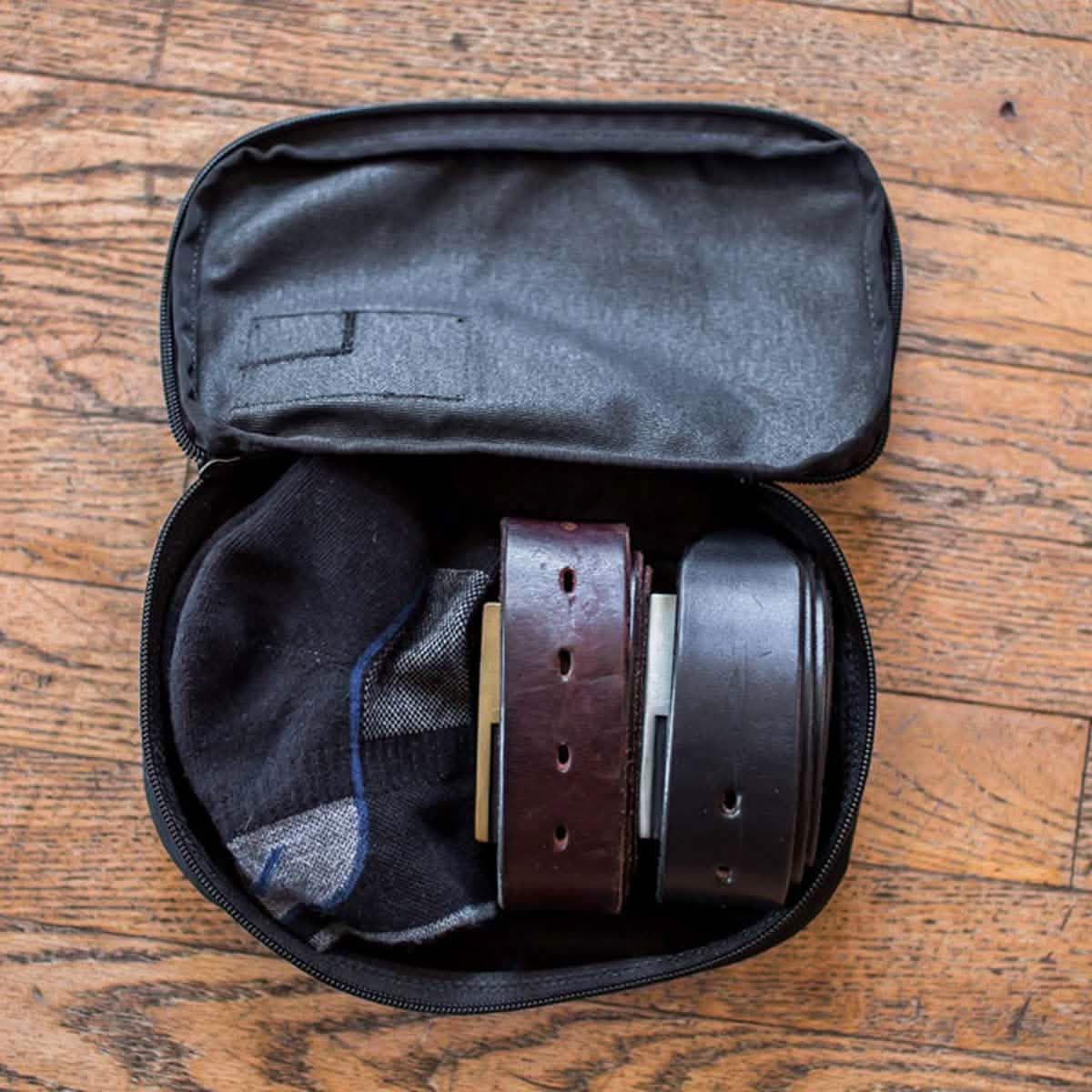Open black travel organizer pouch containing two rolled leather belts and a pair of black socks on wooden floor