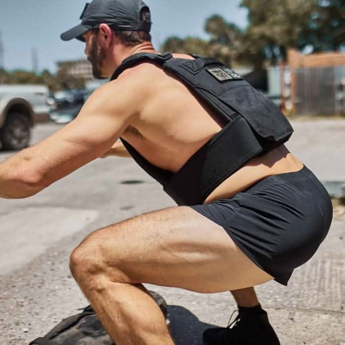 A man sporting a cap, a weighted vest, and durable Men’s Ranger Panties from GORUCK, made with ToughStretch fabric, performs a squat outdoors. In the background, a truck and trees are visible beneath a clear sky.