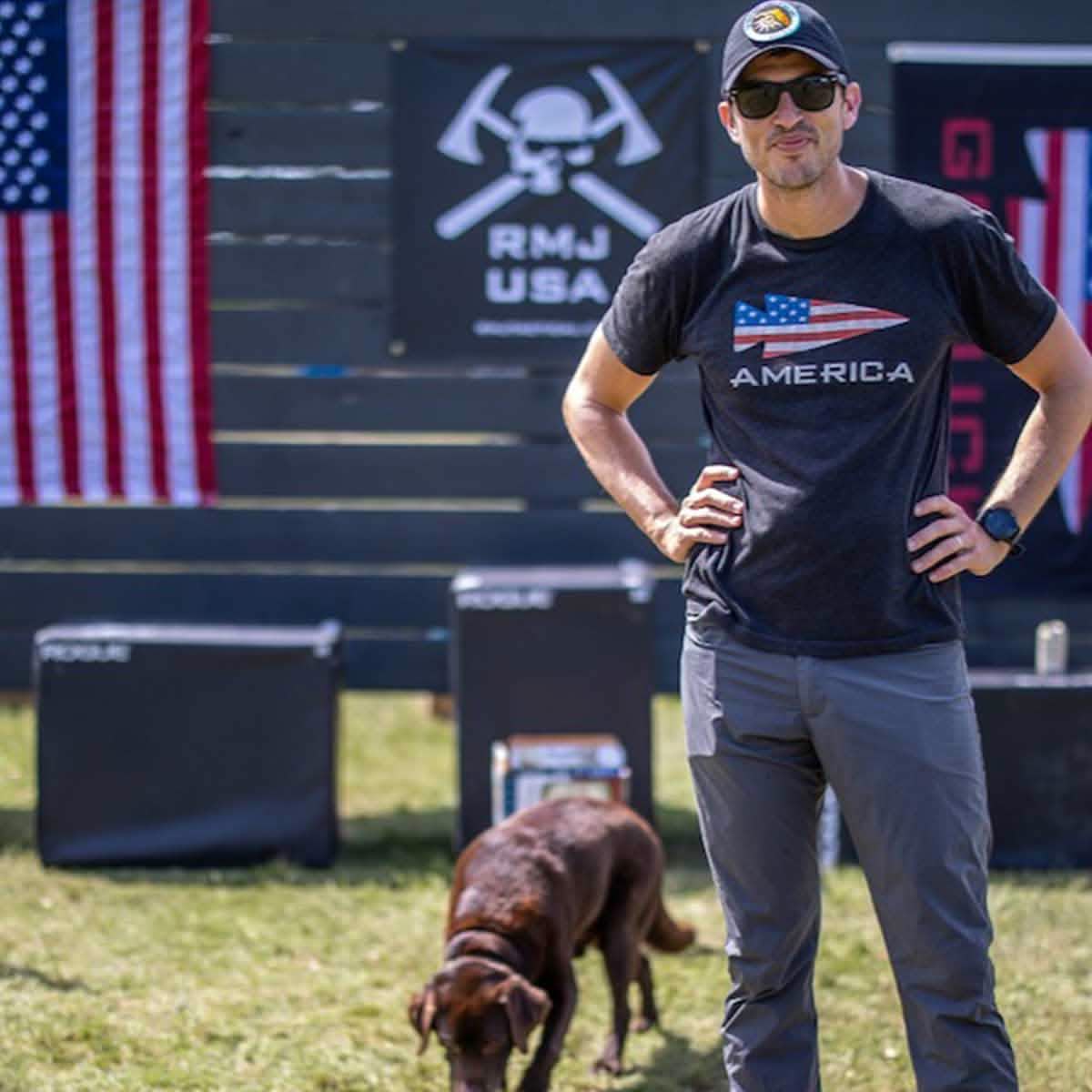 A man in sunglasses stands outdoors wearing a vendor-unknown America Tee - Tri-Blend and a cap. A brown dog sniffs the grass nearby. In the background are American flags and signs, set against a dark wooden fence.