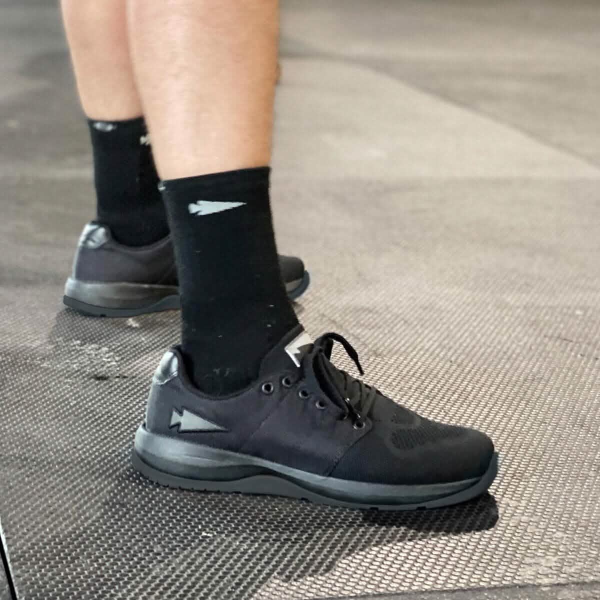 A person stands on a textured floor displaying alternating light and dark sections, ideal for showcasing functional fitness footwear. They are wearing GORUCK's Men's Ballistic Trainers in Blackout, made with CORDURA® Ballistic Nylon and 3X Support™, paired with black socks featuring a white arrow design.