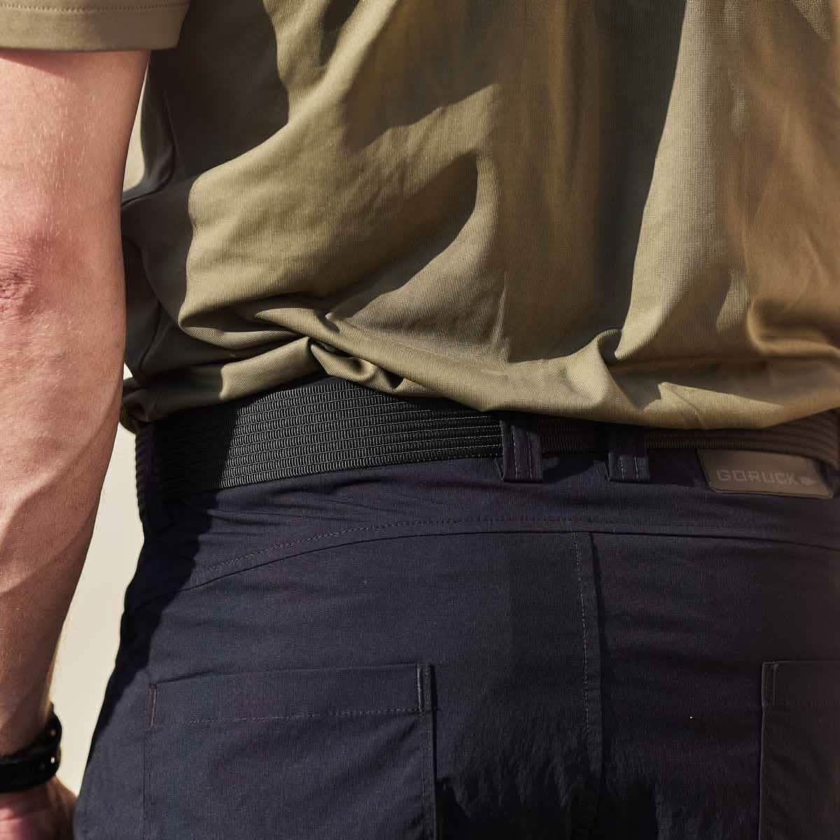 Rear view of a person wearing a green shirt tucked into dark pants, secured with a Grip 6 Spearhead Web Belt featuring a no-slip locking mechanism. The lighting highlights the fabric texture of the shirt and pants, emphasizing the belt's low-profile design.