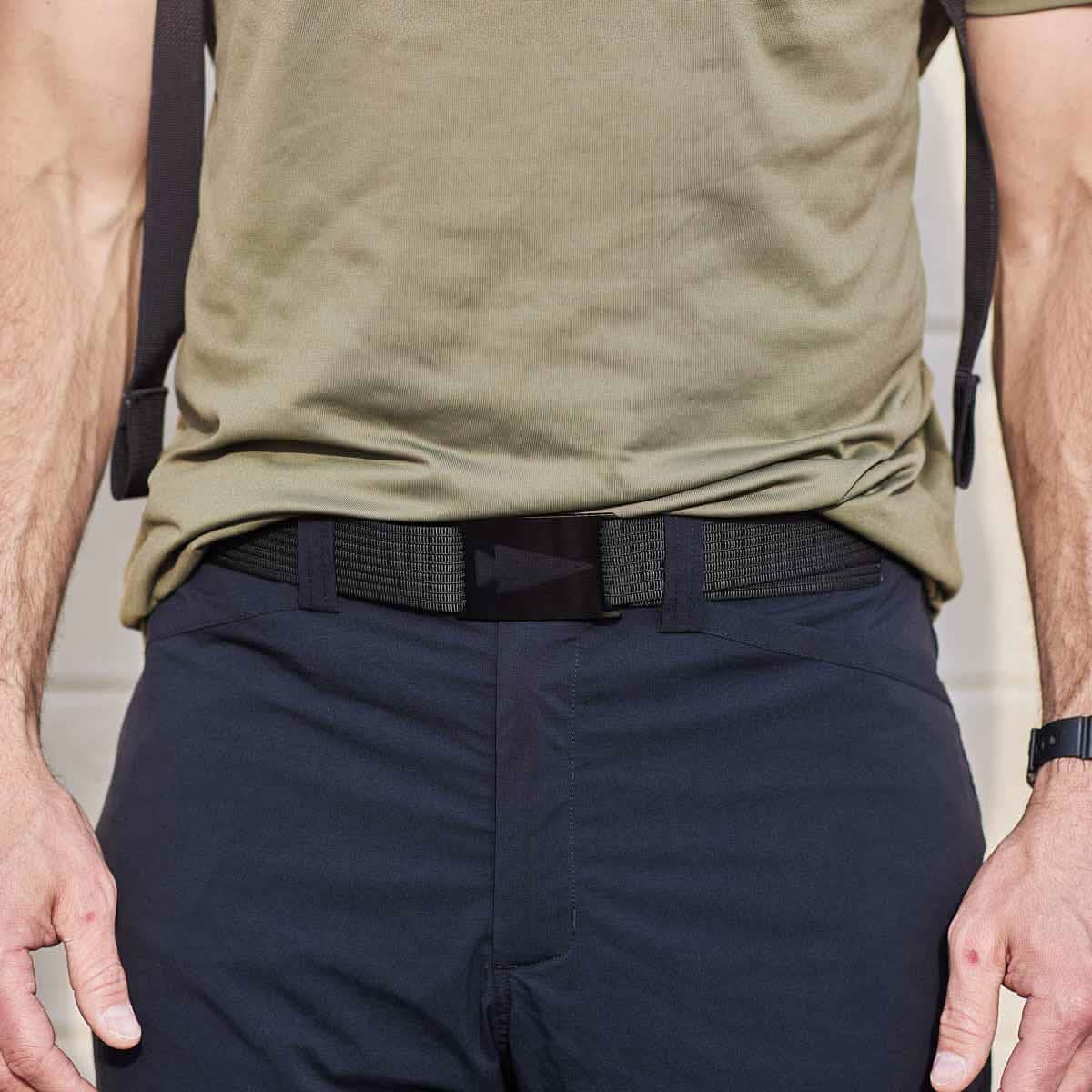 A person is dressed in a green shirt tucked into navy blue pants, accentuated by a black GRIP6 Spearhead Web Belt and suspenders. Their hands rest casually by their sides, emphasizing the belt's sleek design. The photo is cropped at the torso, showcasing the outfit’s polished look.