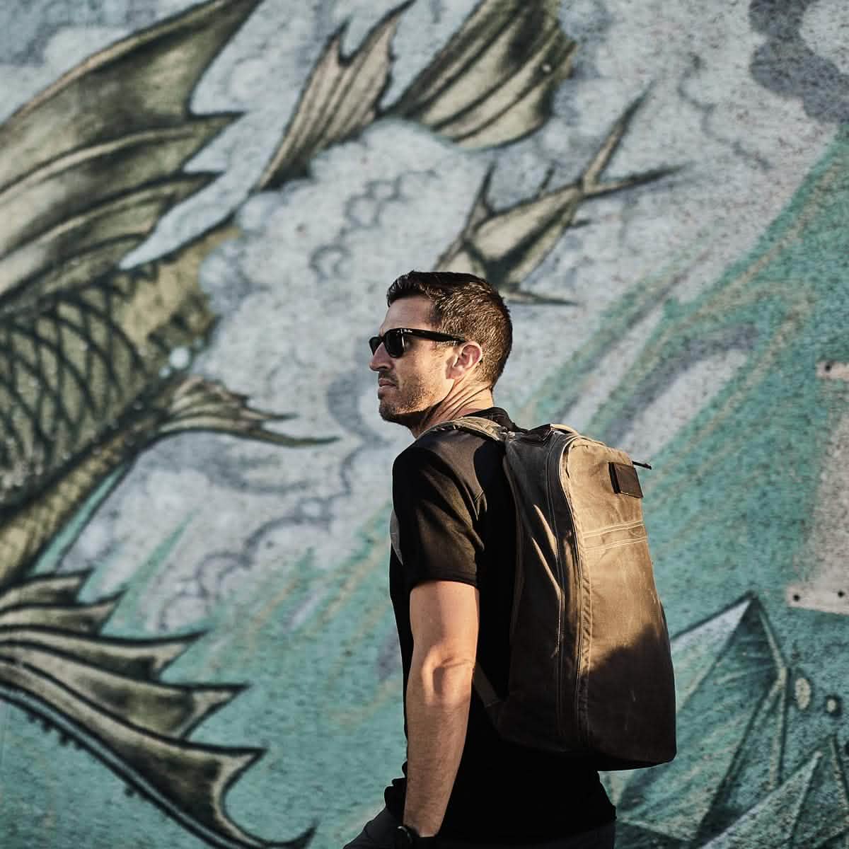 A man wearing a GORUCK Men’s Commando Tee made from merino wool strides in front of a large mural of a fish with dragon-like features. With his brown backpack slung over reinforced paneling, the clear sky casts shadows on the ground.