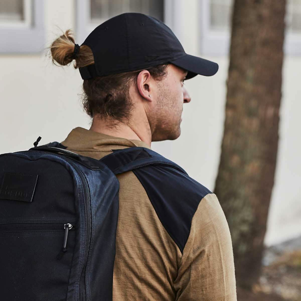 A person with long hair styled in a bun is seen from the side, sporting a black GORUCK Performance TAC Hat - Slick made from TOUGHDRY fabric and a tan jacket. They carry an ample black backpack while standing outdoors near a tree and building, prepared for anything with sweat-wicking efficiency.