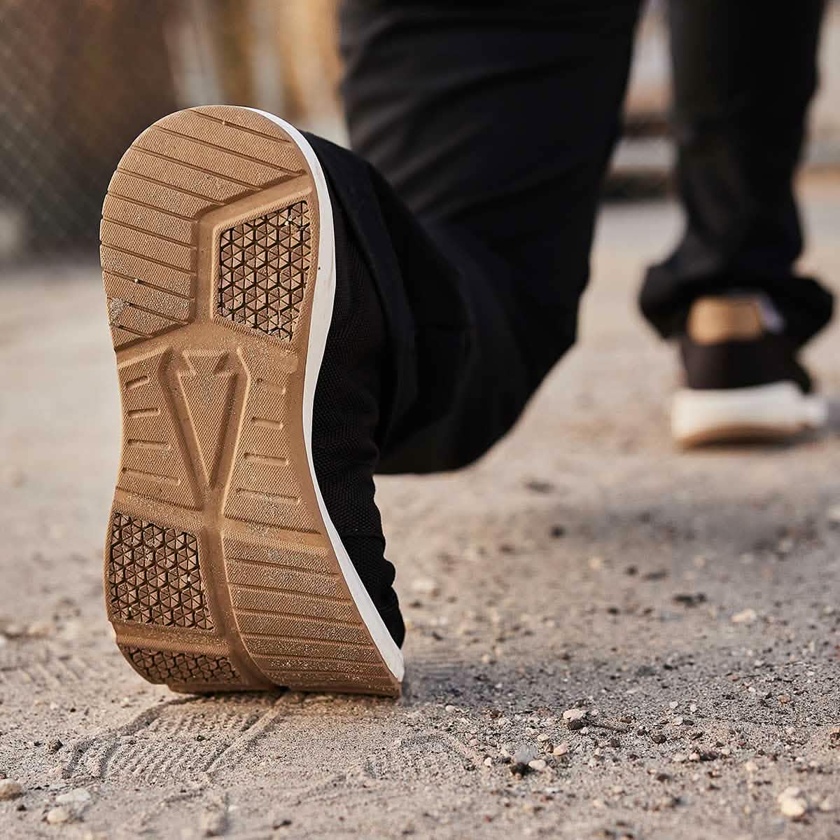 Close-up of a person walking, highlighting the sole of the GORUCK Men's Ballistic Trainers on a sandy surface. The shoe's geometric pattern is striking, crafted with CORDURA® Ballistic Nylon. The slightly blurred background reveals hints of a fence.