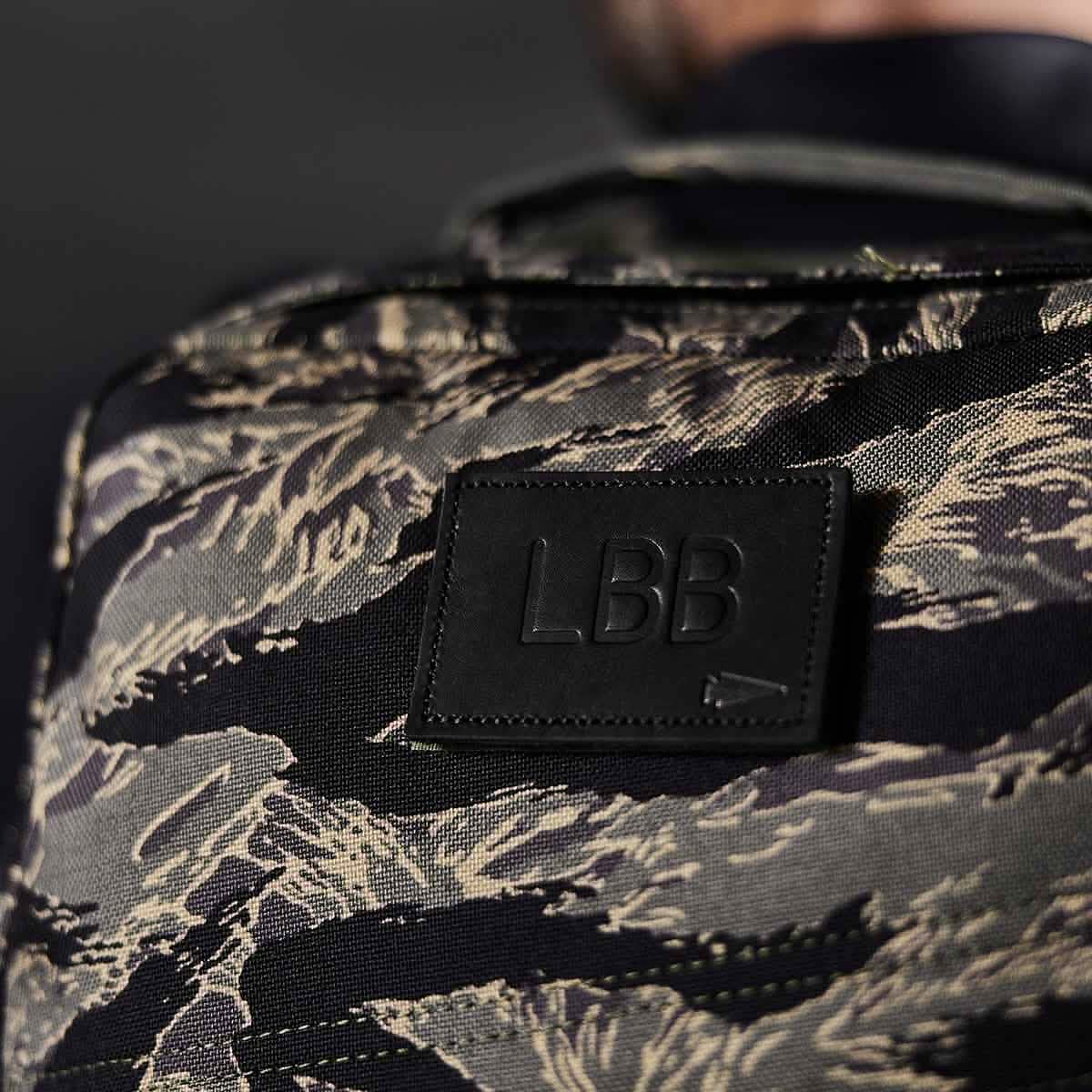 A close-up of a camouflage-patterned backpack highlights the Patch - Custom Leather Monogram by vendor-unknown, showcasing the black initials LBB. The dark background brings out the backpack's textured fabric and personalized style.