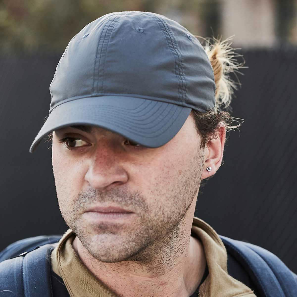 A person wearing a GORUCK Performance TAC Hat - Slick - TOUGHDRY and a brown jacket looks to the side. The individual, sporting a backpack and an earring, has their hair tied back. The background is blurred, emphasizing the person's contemplative expression.