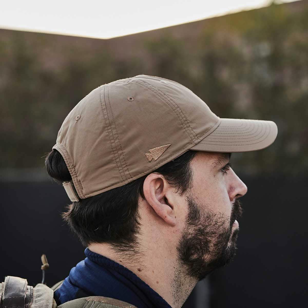 Side view of a person wearing the beige Performance TAC Hat - Slick by GORUCK outdoors, facing right. Made from TOUGHDRY fabric, the hat features a small logo on the side. The background is blurred, suggesting trees or bushes. This sweat-wicking performance hat perfectly complements the person's beard and dark jacket.