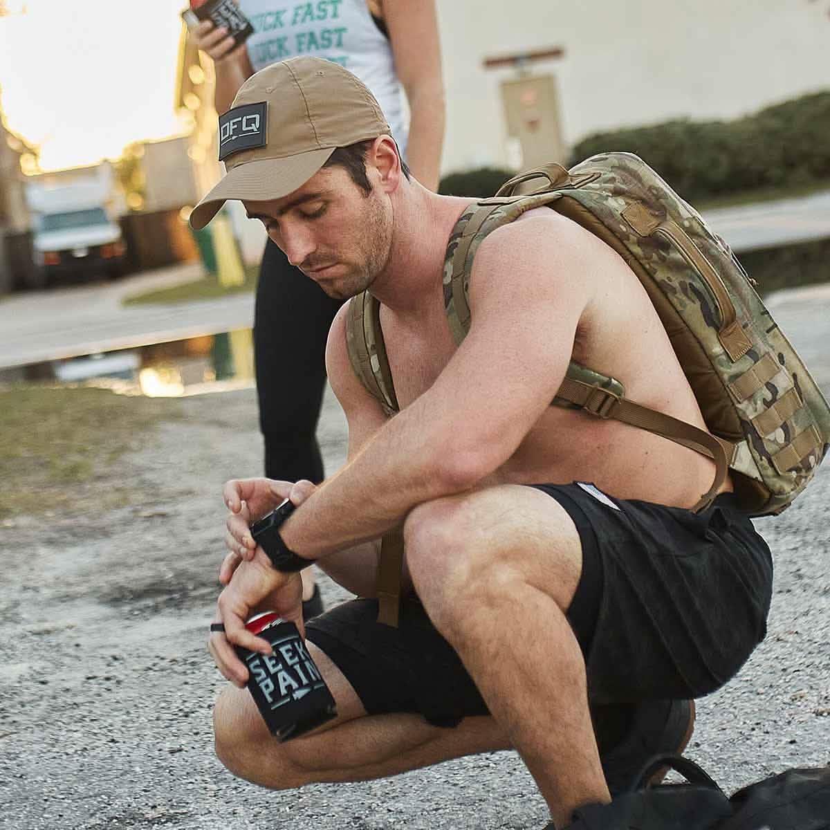Man wearing GORUCK coyote tactical hat and backpack, crouching outdoors holding drink can