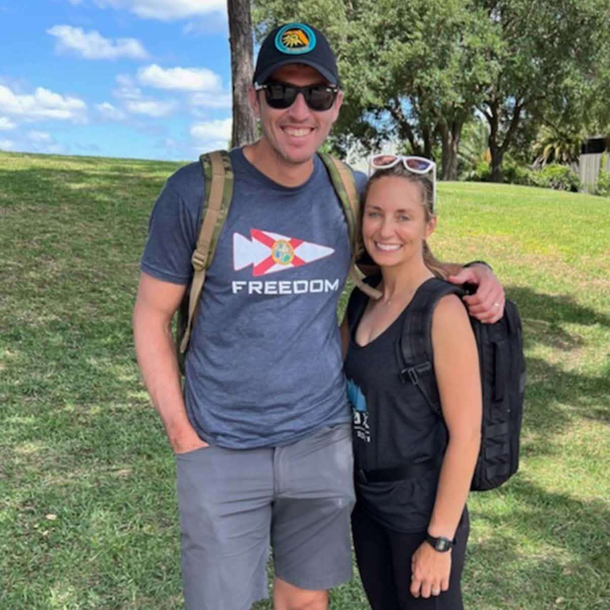 Two people outdoors wearing GORUCK rucksacks, smiling on grass under trees, casual attire