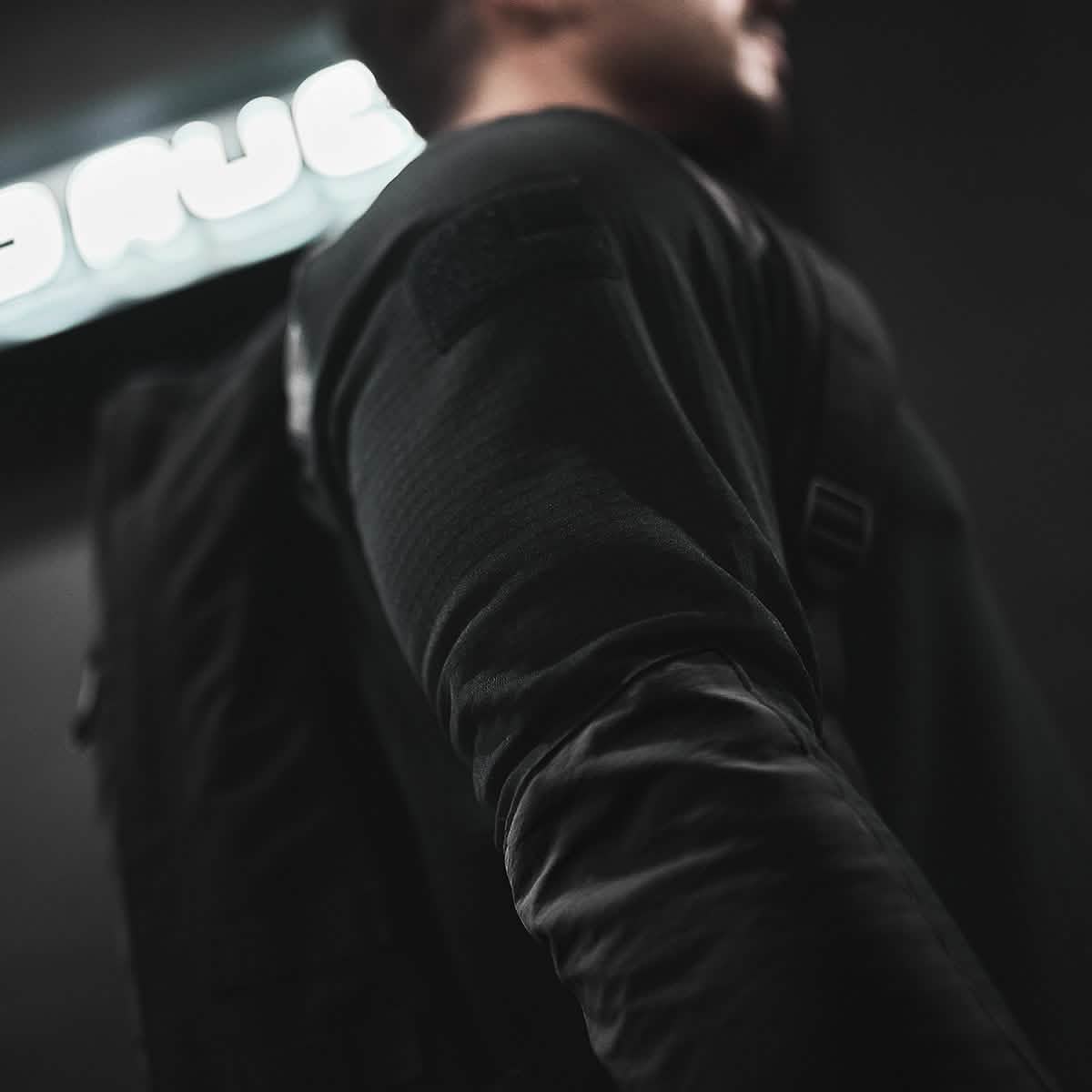 A person wearing the black Men’s Half Zip - Polartec Grid Fleece by GORUCK, along with a backpack, stands in a dimly lit setting. The emphasis is on their arm and shoulder, while a blurred neon sign softly illuminates the Polartec Power Grid fabric in the background.
