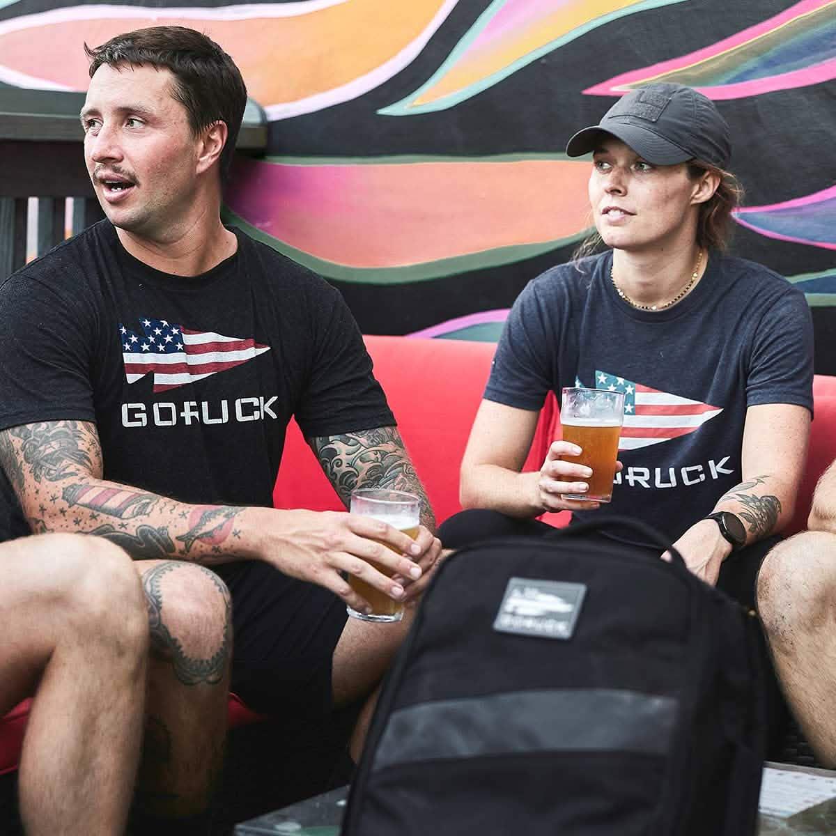 Two people wearing black GORUCK t-shirts with American flag design, sitting on a red couch holding beers with a colorful mural behind