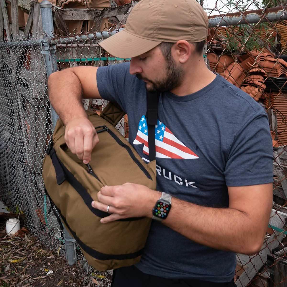 Man wearing tan cap and GORUCK USA t-shirt opening brown GORUCK bag near chain-link fence with stacked tiles