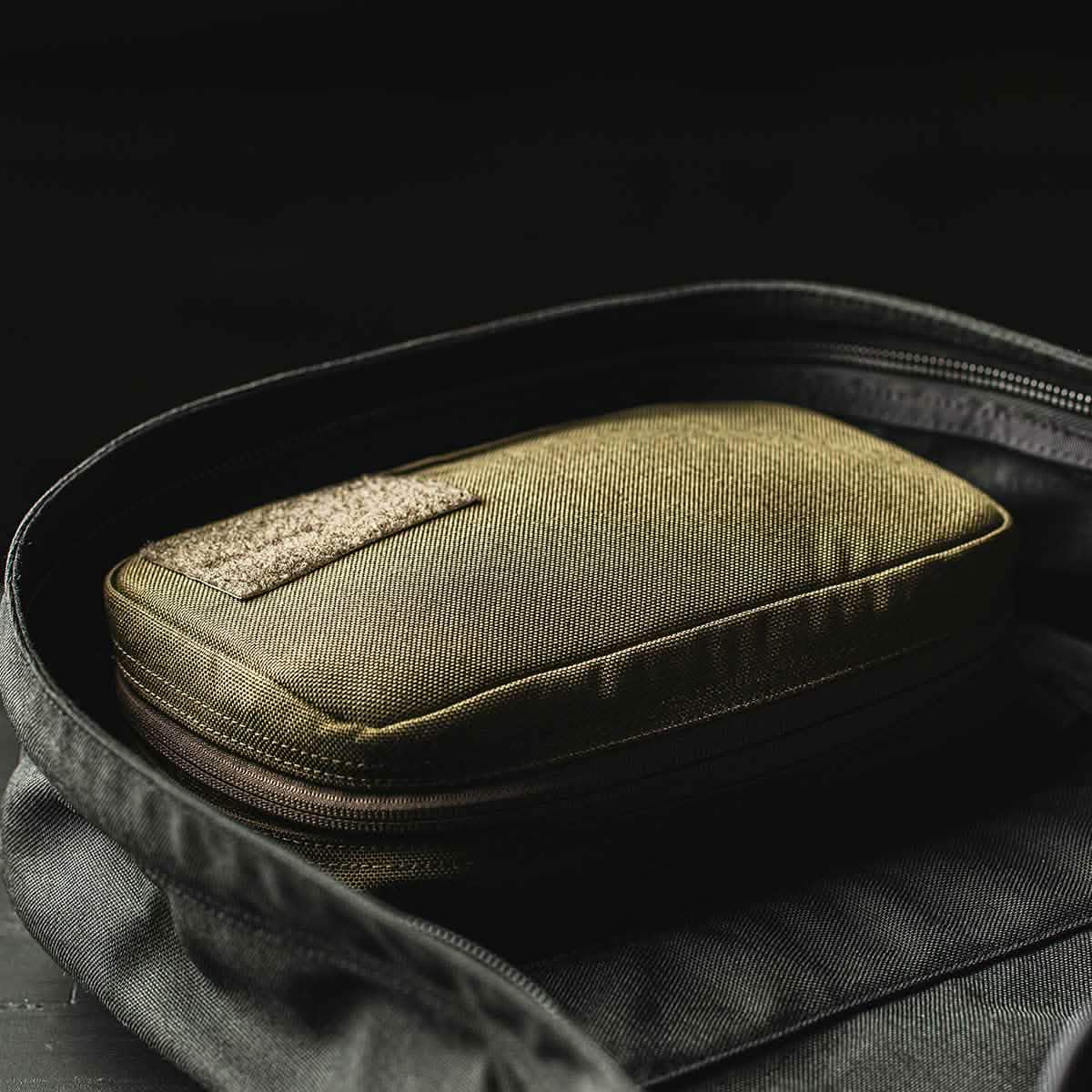 A GR1 Field Pocket from GORUCK, featuring a textured khaki design and a rectangular patch on top, is nestled inside an open black bag. The surrounding bag is unzipped, showcasing the neatly positioned pocket.