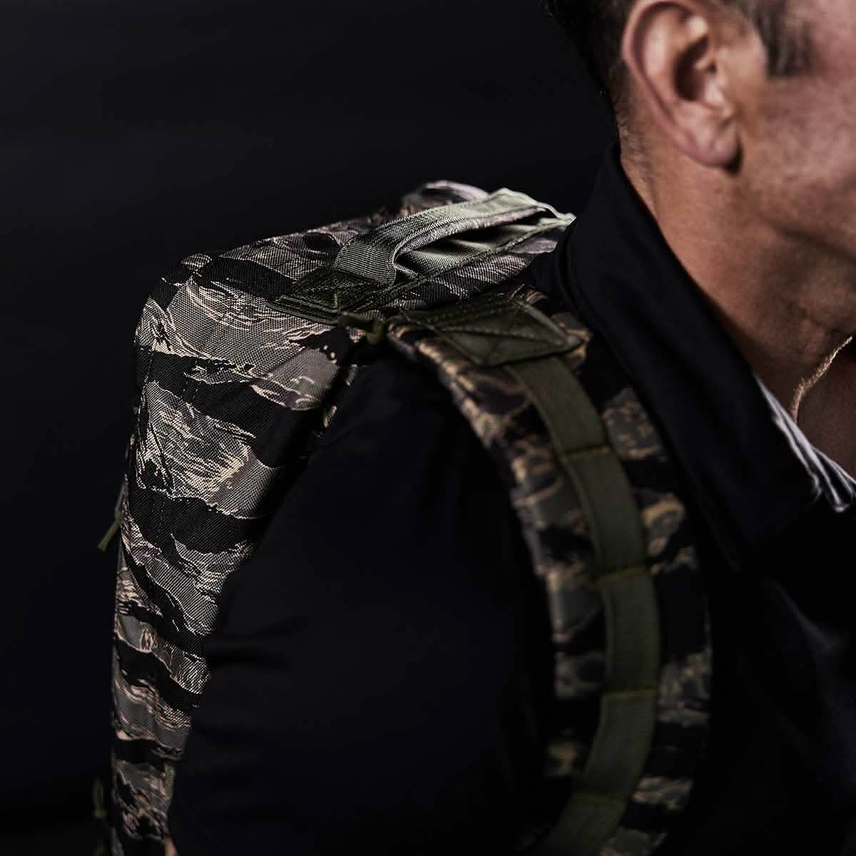 Close-up of a person wearing the GR1 - USA - Tiger Stripe rucksack, featuring a bold camouflage pattern inspired by the iconic Special Forces backpack, against a dark background.