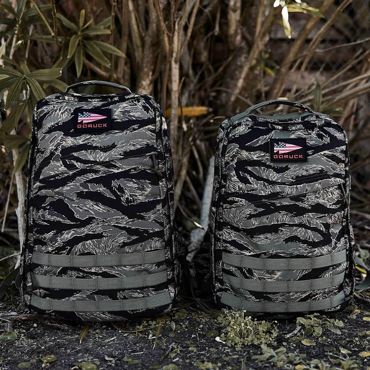 Two GR1 - USA - Tiger Stripe rucksacks stand side by side outdoors, highlighting American manufacturing excellence with lush plants and trees in the background.