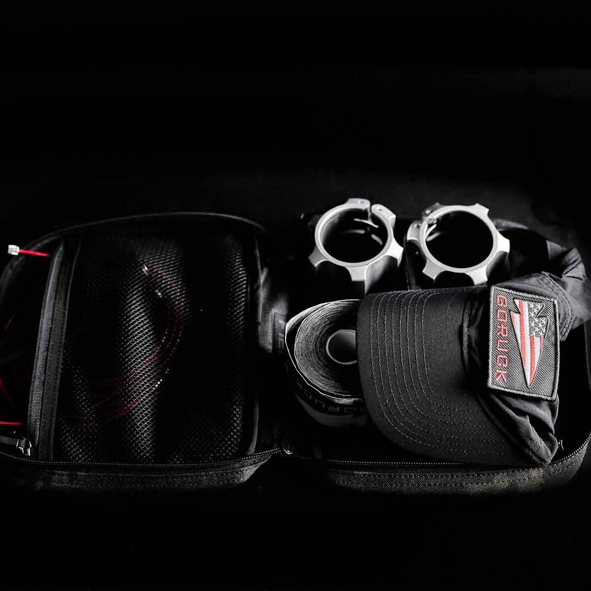 Open the GR2 Field Pocket - Black Multicam to find an electronics organizer, two silver metal objects, a rolled-up strip, and a black GORUCK baseball cap.