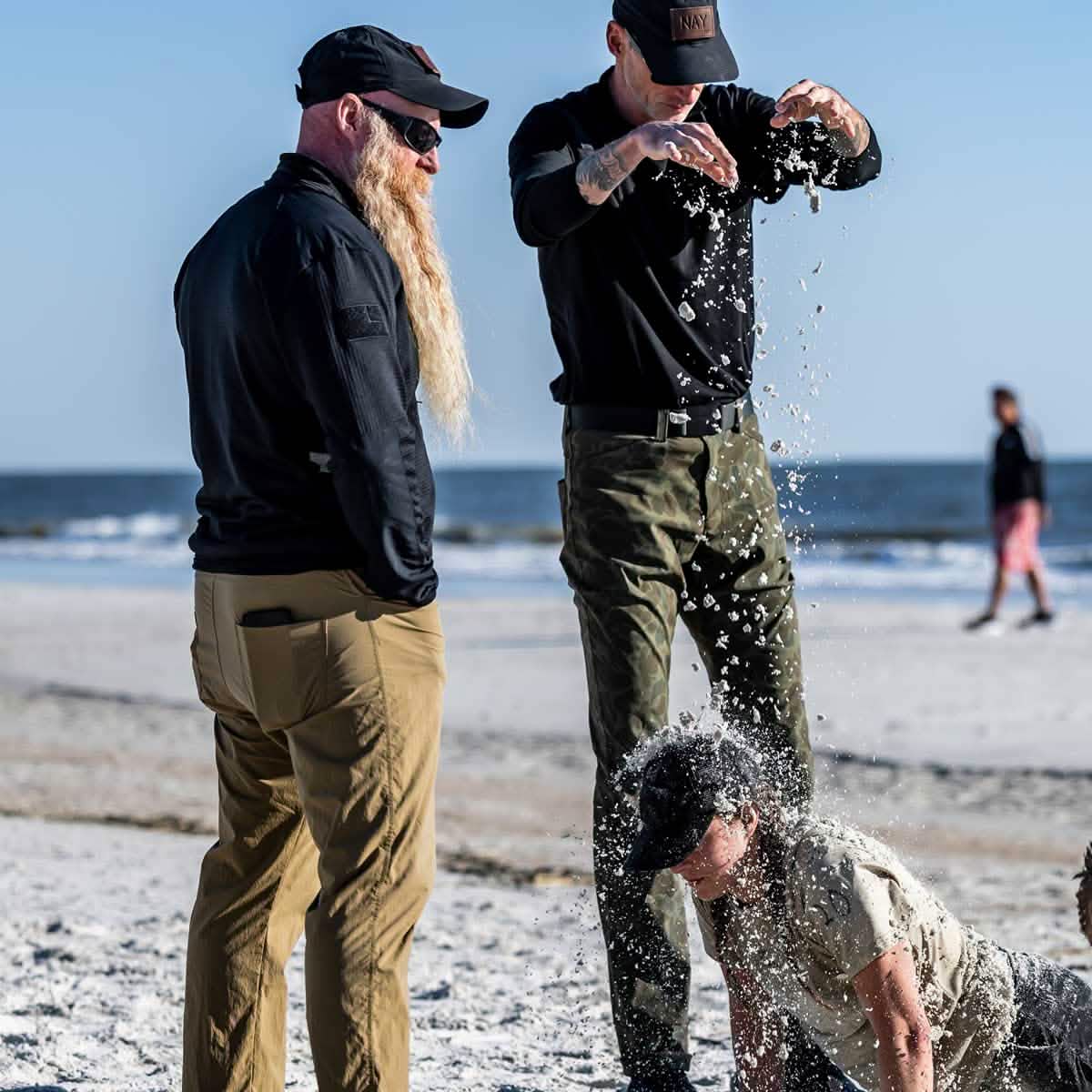 Three people on a beach: one with a long beard standing in the Men's Half Zip - Polartec Grid Fleece by GORUCK, another pouring sand, and a person doing push-ups on the ToughDry fabric towel. The ocean is in the background, with a few others walking along the shore.