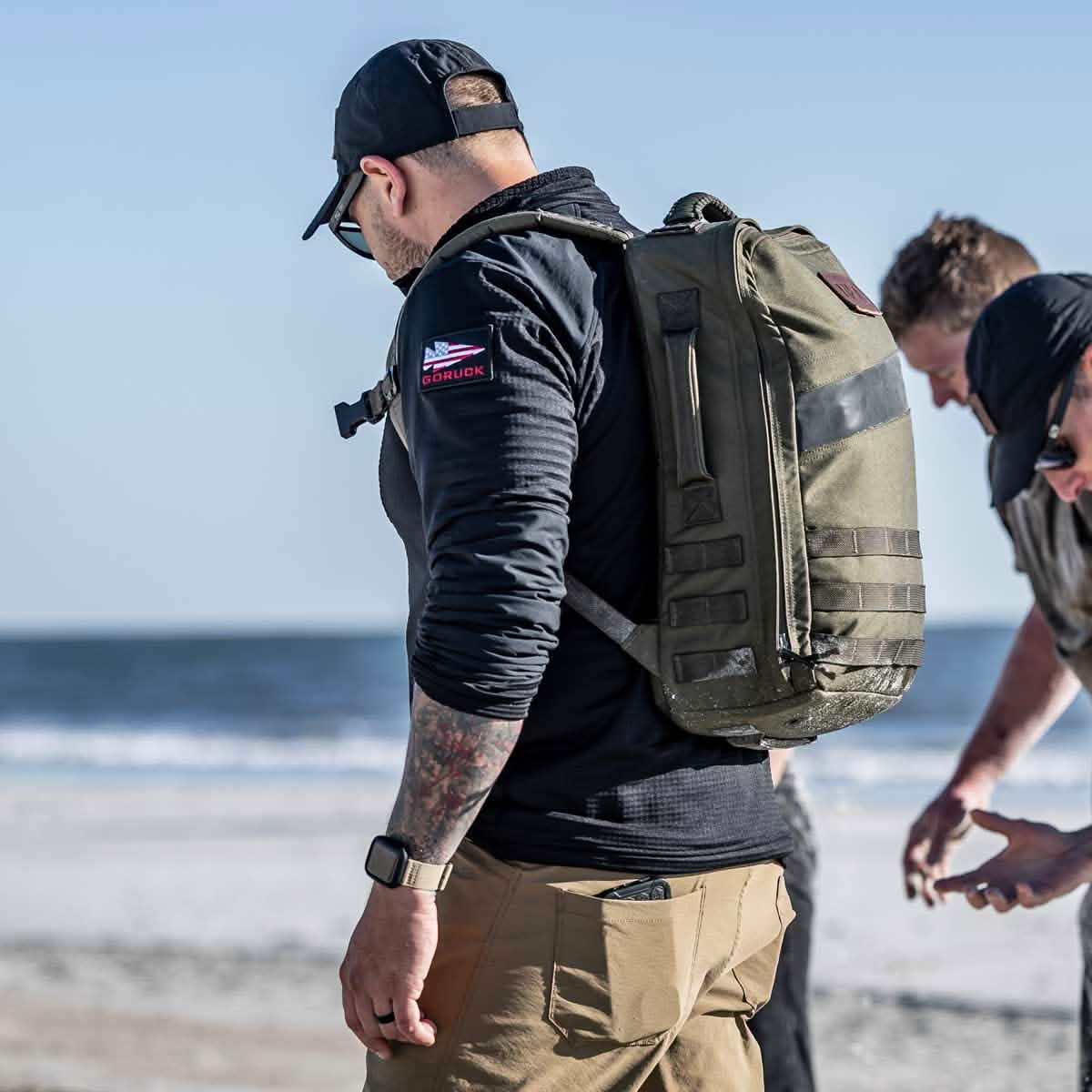 A man, donned in a Men’s Half Zip by GORUCK with a patch on the arm and wearing a black cap and sunglasses, carries a large green backpack made from ToughDry Fabric. Beside him, another person crouches slightly while gazing at the ocean in the background.