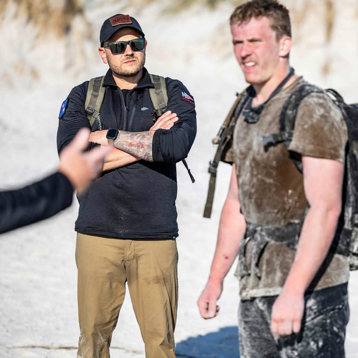 Two men on a beach, both donning GORUCK tactical attire with one wearing the Men's Half Zip - Polartec Grid Fleece. The man on the left stands confidently with arms crossed, sporting sunglasses and a cap. The man on the right is covered in sand, gazing ahead. An arm of another person appears in the foreground.