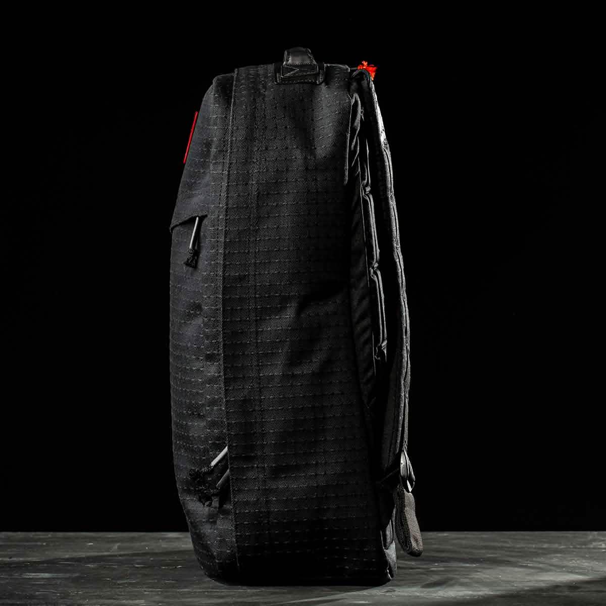 The GR1 USA x Carryology - GRXC2 Samurai backpack, made from Japanese cotton, stands upright on a dark surface against a black background, displayed in profile.