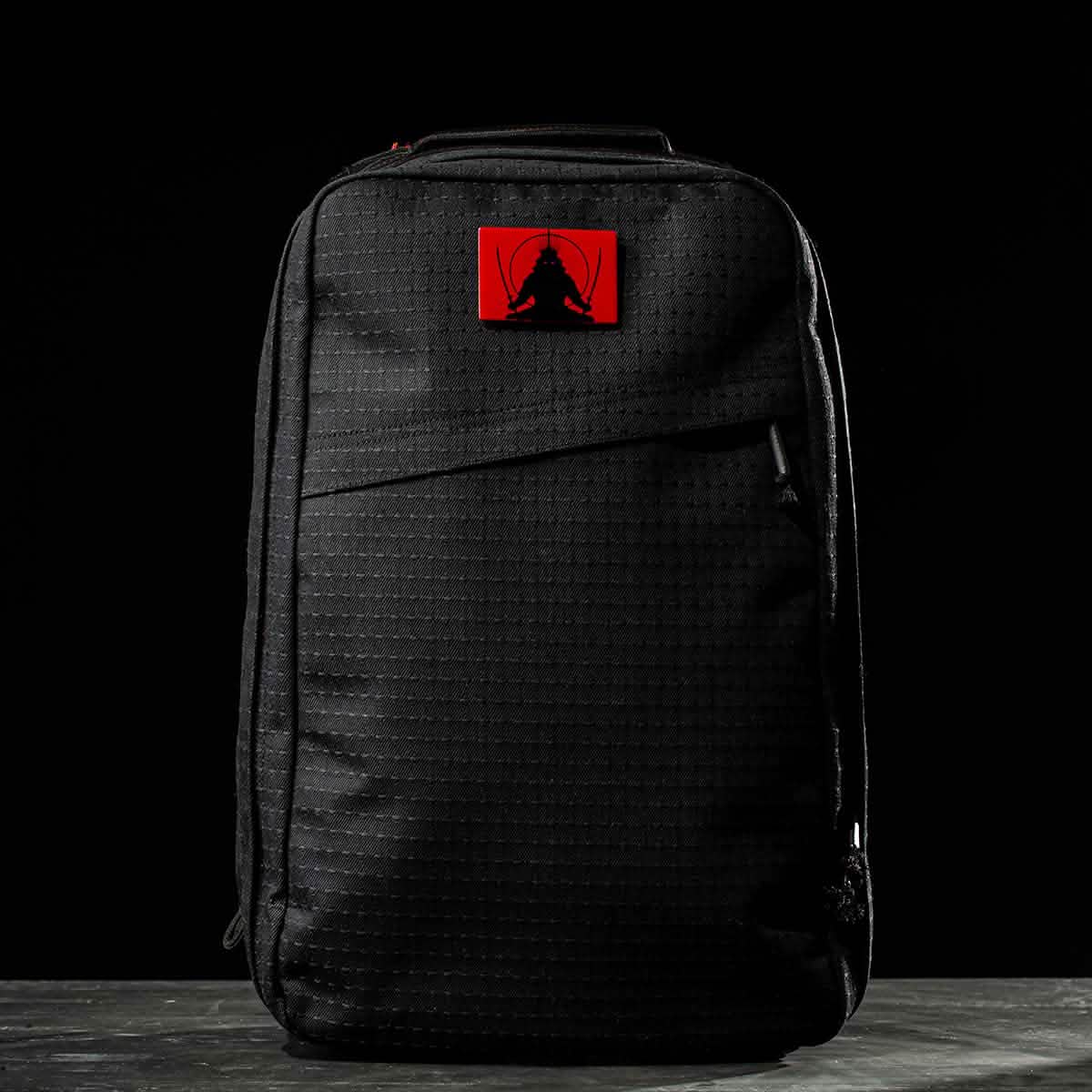A black GR1 USA x Carryology - GRXC2 Samurai backpack made from Japanese cotton, featuring a red patch with a black abstract design. Part of the exclusive Samurai GR1 collection.