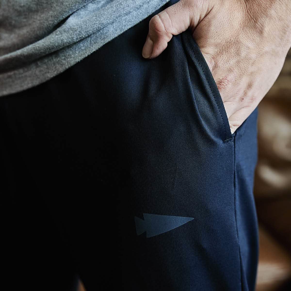 Close-up of a person's hand in the pocket of dark blue Indestructible Performance Joggers by GORUCK, made from moisture-wicking fabric and showcasing a small arrowhead logo. They are paired with a gray top.