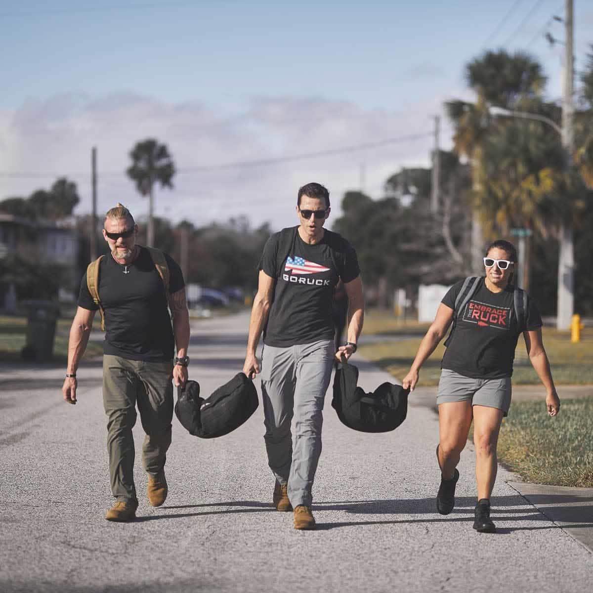 Three people walking on a sunny street carrying GORUCK weighted bags, wearing GORUCK apparel and sunglasses