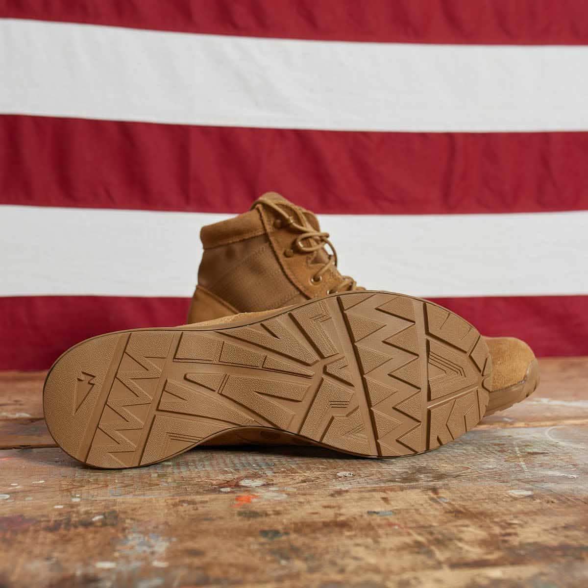 A MACV-1 - Mid Top - Coyote Suede work boot by GORUCK crafted from suede leather lies on its side with the sole up, highlighting its all-terrain outsole and tread pattern. It rests on a wooden surface against a backdrop of red and white striped fabric.